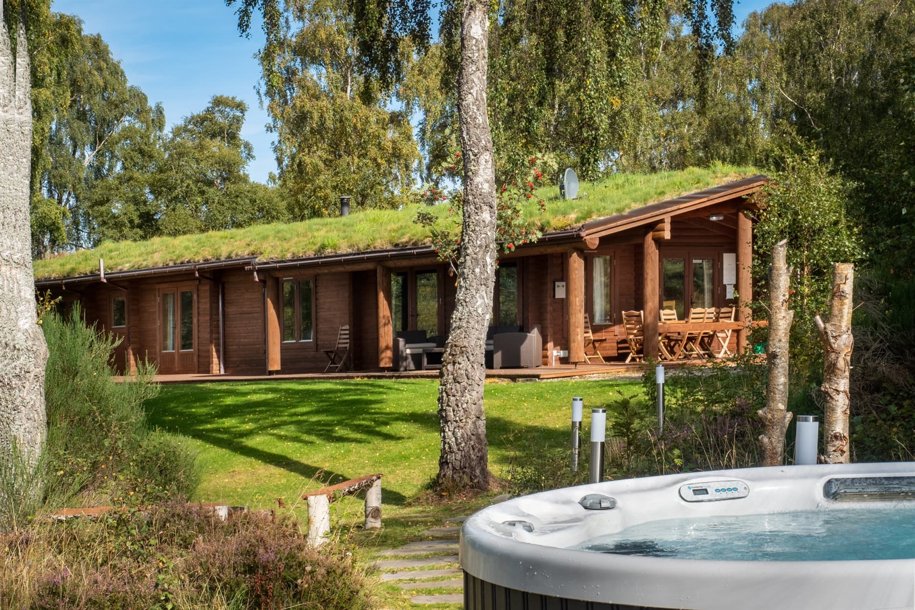View of the log cabin from the garden. The hot tub is in the foreground and there is a table on the deck area in front of the lodge.