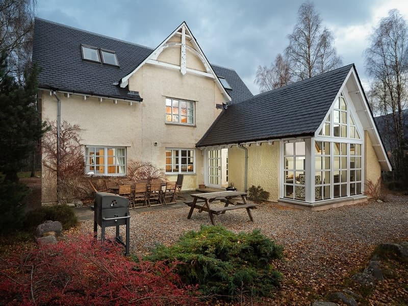 There is a large house with many window. Outside the house is a gravel patio area a dining table and chairs and a picnic bench.