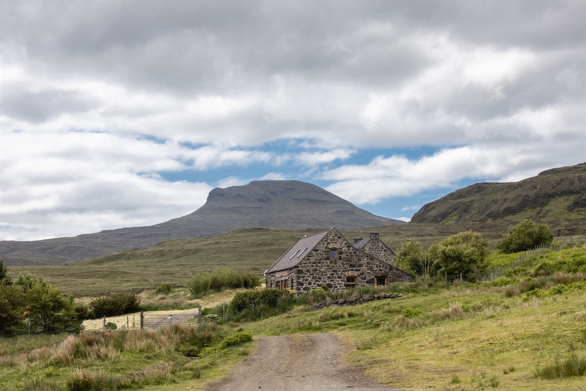 The view from the farm track towards the stone built cottages with Macleod's Tables in the background and a blue and cloudy sky