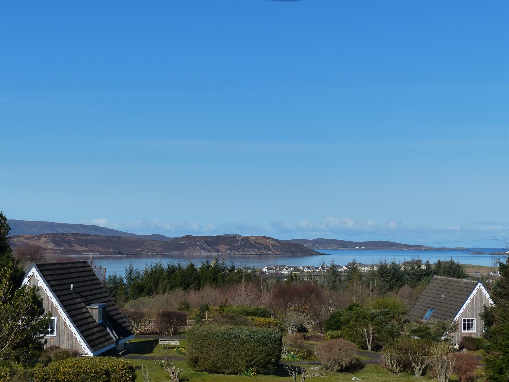 Aultbea Lodges, 2 of our 5 larch clad A-Frame Highland Lodges, lodge grounds with trees and bushes, Loch Ewe and the Isle of Ewe in the background wit