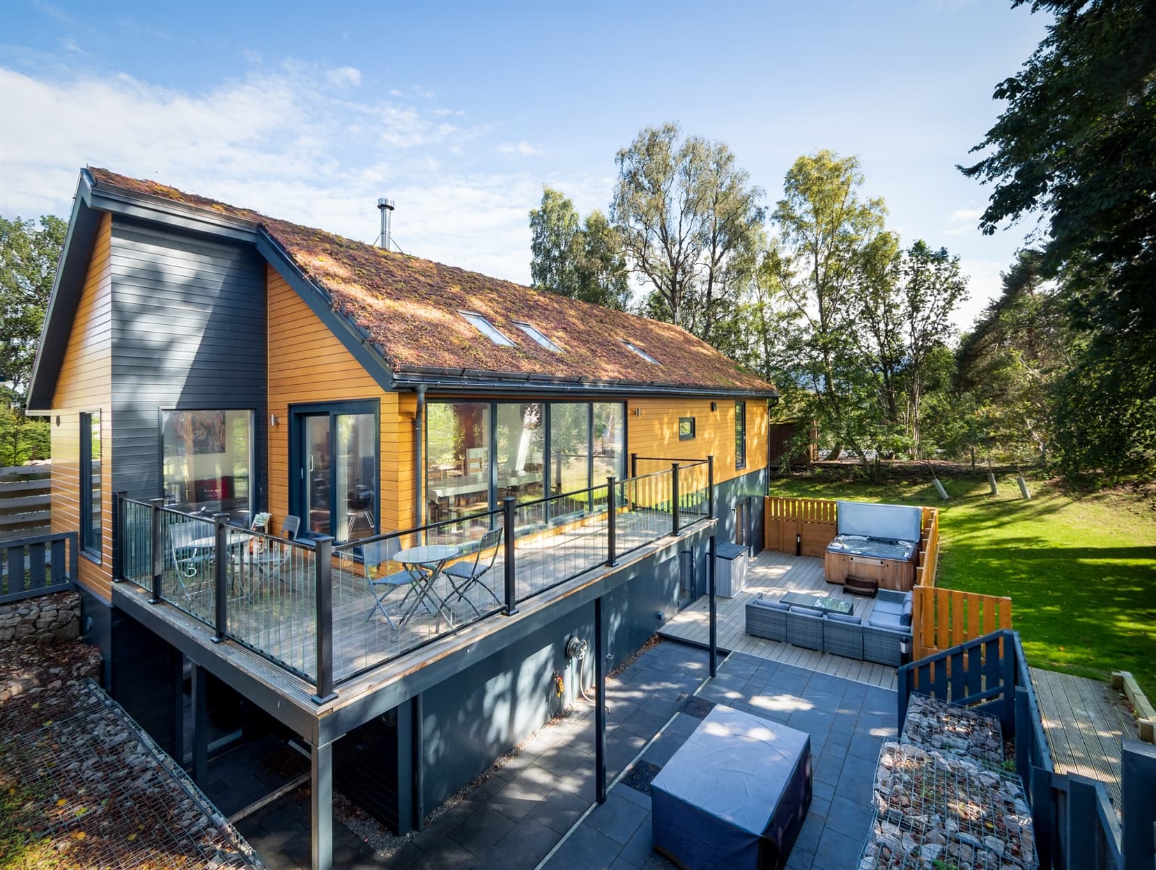 An exterior shot showing the lodge. There is a large warp around balcony on the upper level and a paved patio area with hot tub and outdoor furniture