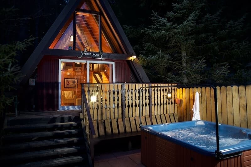 Looking at the exterior of the lodge from the garden at night time. The lodge is an A frame timber cabin with a hot tub in front of it.