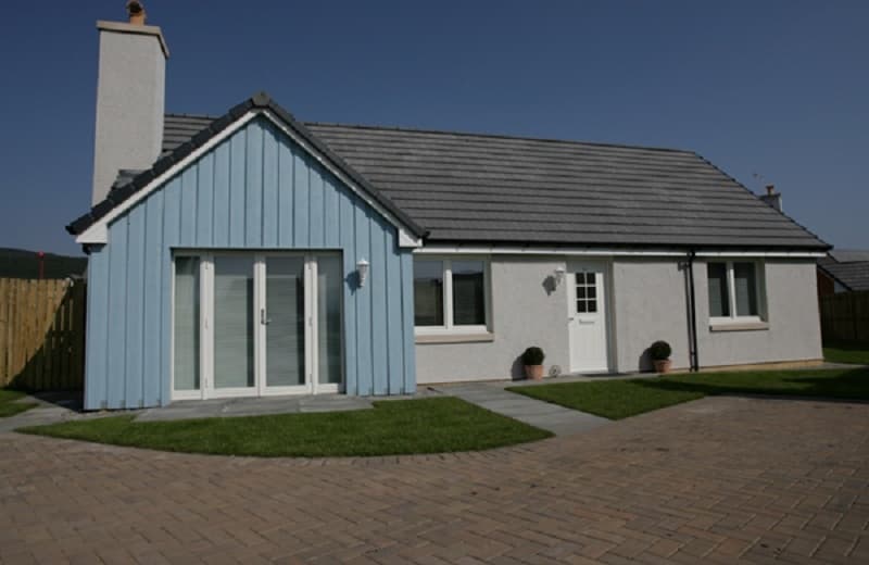 View of Heather Cottage from the road. It is a modern bungalow with blue timber cladding. There is a paved driveway.