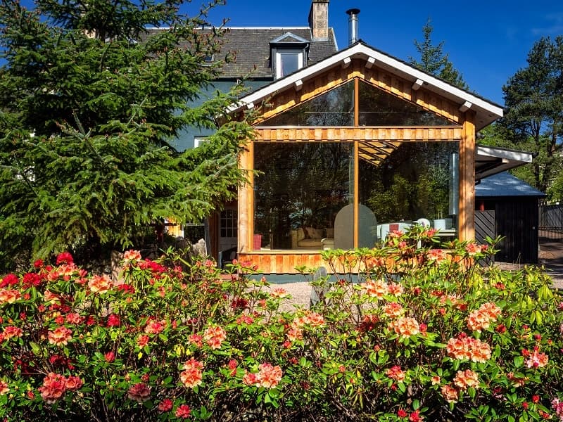 View from the garden looking towards the house. There are orange and red flowers on the bushes. The house has timber cladding and large windows.