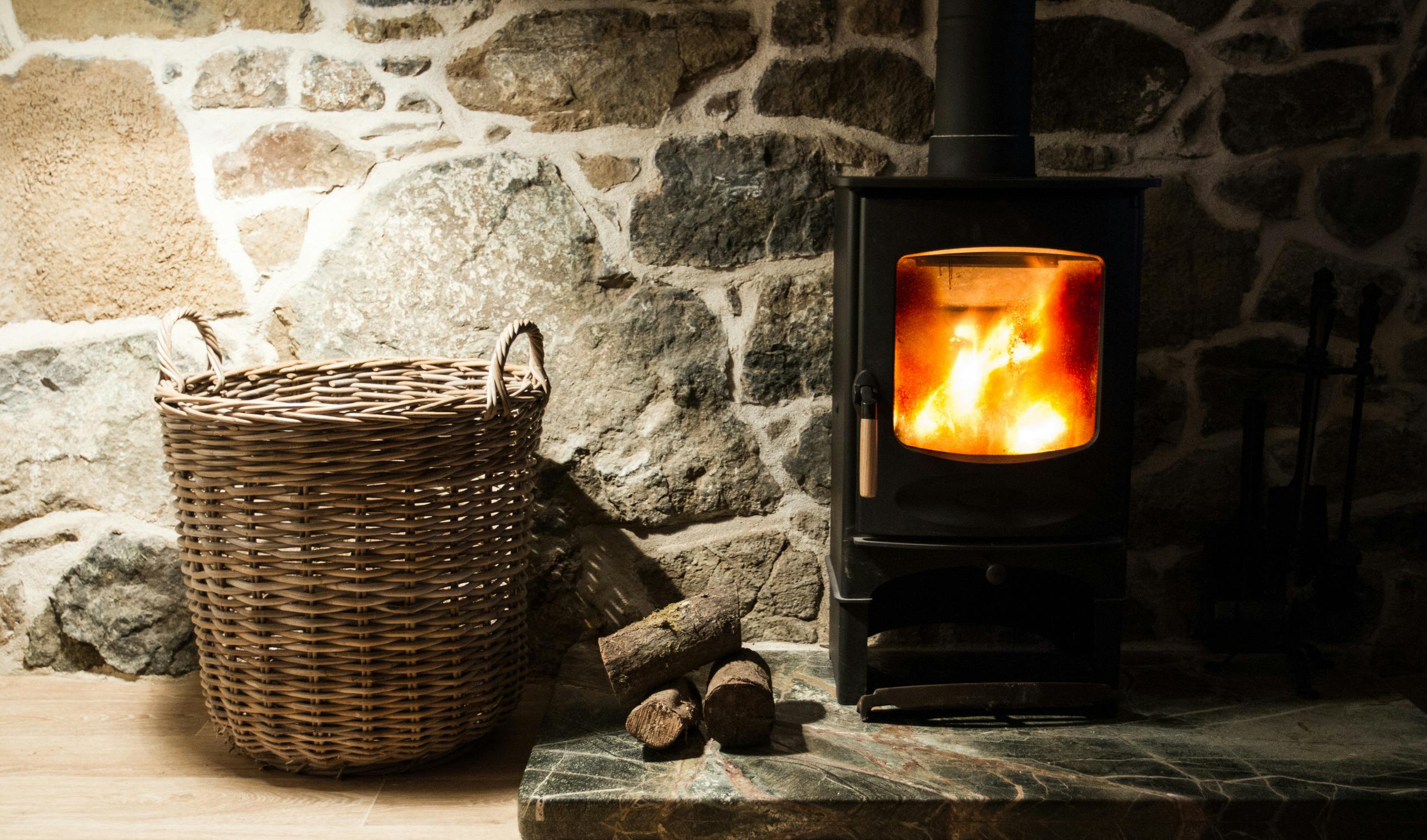 A wood burner drive and fireplace inside an old. stone cottage with logs and wicker storage basket.